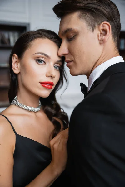 Woman with red lips looking at camera near boyfriend in suit at home — Stock Photo