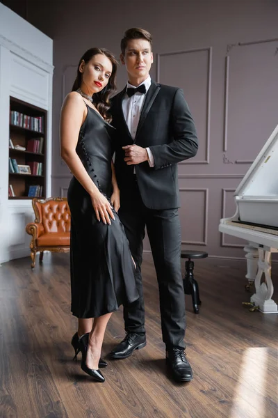 Stylish woman in silk dress and heels looking at camera near elegant boyfriend at home — Stock Photo