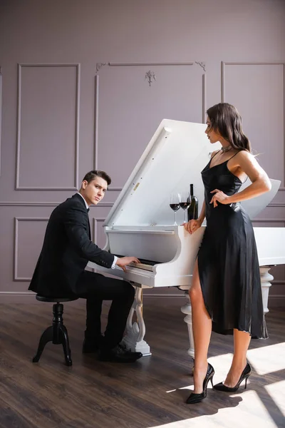 Elegant man looking at camera while playing piano near girlfriend and wine at home — Stock Photo