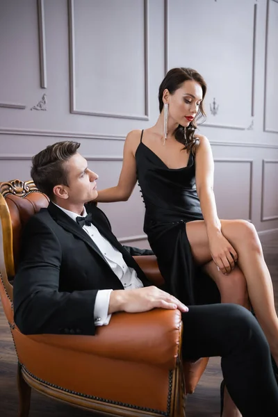 Elegant man looking at sensual girlfriend while sitting on armchair at home — Stockfoto