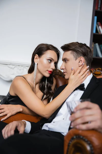 Sensual woman in silk dress touching boyfriend in suit holding blurred glass at home — Stock Photo