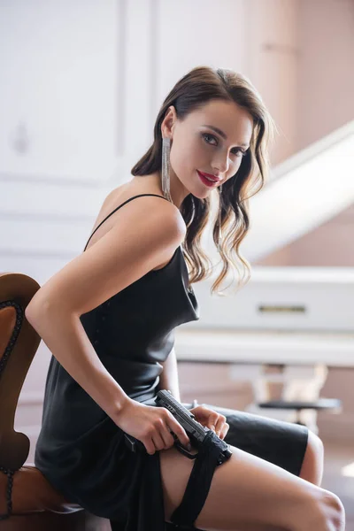 Smiling woman in dress putting gun in garter and looking at camera on armchair — Stock Photo