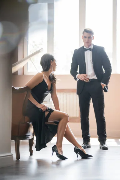 Sensual woman in heels and dress putting handgun in garter near boyfriend with whiskey at home — Stock Photo