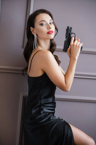 Brunette woman in silk dress holding handgun and looking at camera near wall — Stock Photo