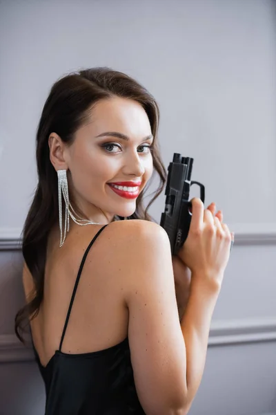 Smiling woman with red lips holding handgun and looking at camera near wall - foto de stock