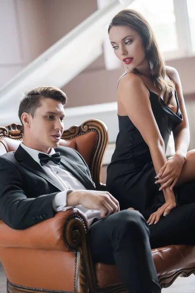 Sensual woman in silk dress touching elegant boyfriend while sitting on armchair — Stockfoto