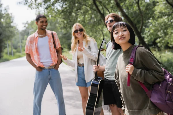 Asian woman with backpack looking away near blurred interracial friends on road — Fotografia de Stock