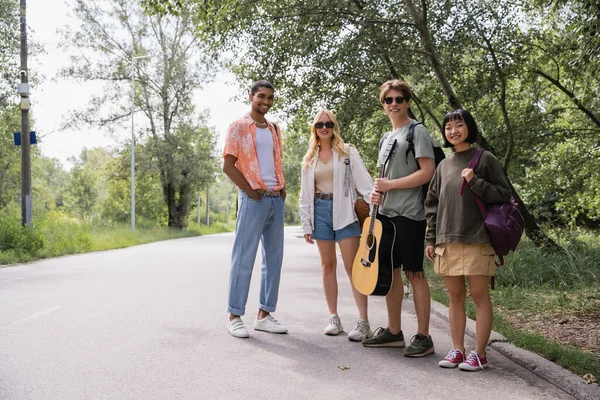 Man with guitar and multiethnic friends with backpacks on road near forest — Fotografia de Stock