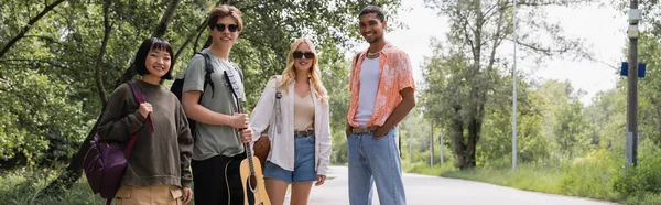 Happy interracial friends with guitar and backpacks standing on countryside road, banner — Foto stock