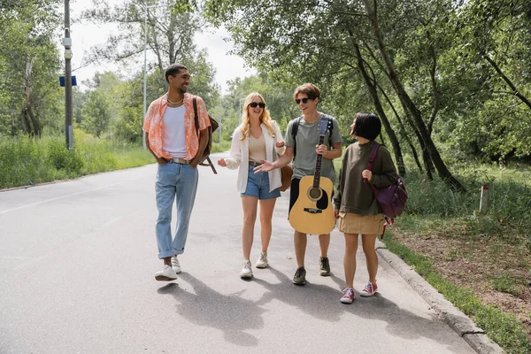 Человек с гитарным жестом и беседой с многонациональными друзьями во время прогулки по дороге — стоковое фото
