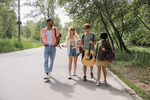 Man with guitar gesturing and talking to multiethnic friends while walking on road — Fotografia de Stock