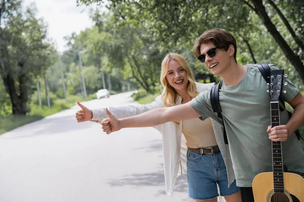 Cheerful hitchhikers with acoustic guitar stopping car with thumbs up — Stock Photo