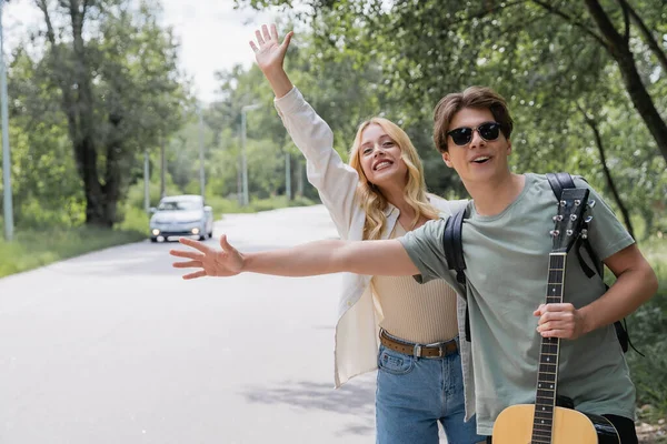 Smiling couple waving hands and stopping car while hitchhiking in countryside — Stockfoto