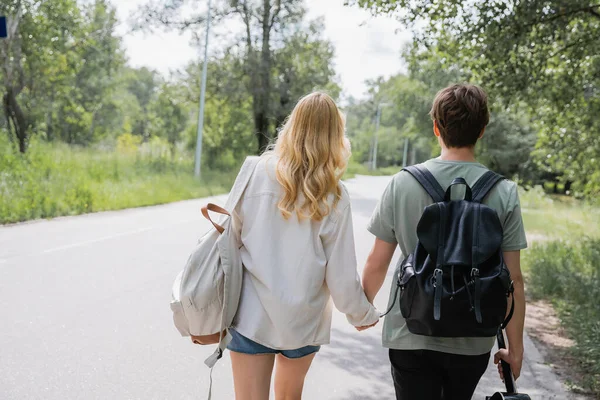 Back view of young travelers holding hands and walking on road along forest — Stock Photo