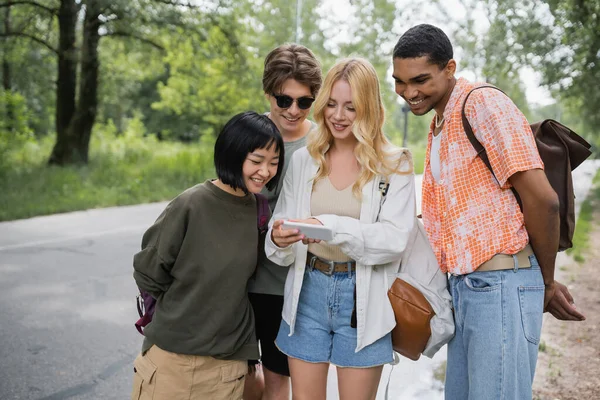 Blonde woman searching location on smartphone near cheerful multiethnic friends — Fotografia de Stock