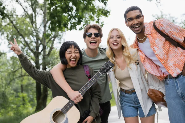 Excited asian woman laughing near happy interracial friends with guitar and backpacks — Stock Photo