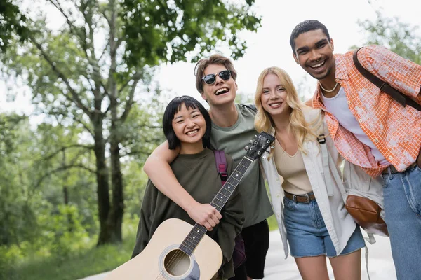 Happy multiethnic travelers with acoustic guitar and backpacks smiling at camera in countryside — Photo de stock