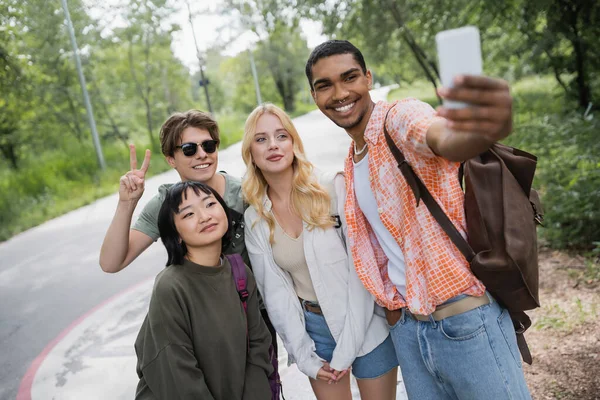Чоловік в сонцезахисних окулярах показує знак перемоги біля міжрасових друзів і афроамериканця, який приймає селфі на розмитому смартфоні — стокове фото