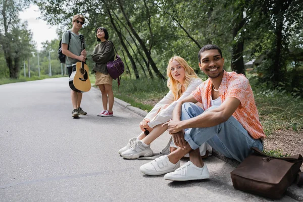 Smiling interracial travelers sitting on road near blurred friends with guitar — Stock Photo