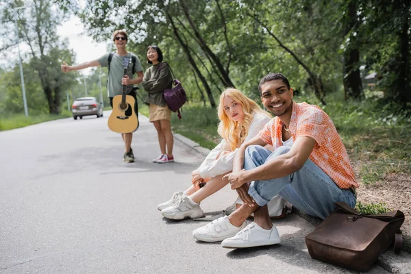 Blurred hitchhikers stopping car near smiling interracial friends sitting on road — Photo de stock