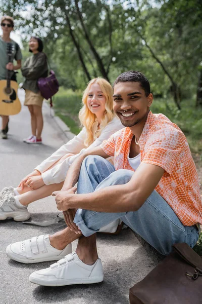 Happy african american man smiling at camera while sitting on road near interracial friends — Photo de stock
