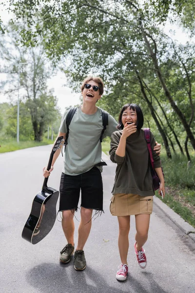 Excited interracial couple with backpacks and guitar laughing while walking on road - foto de stock