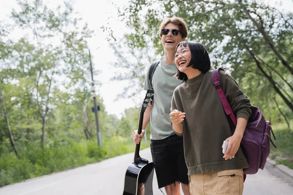 Excited asian woman laughing near boyfriend in sunglasses while walking in countryside - foto de stock