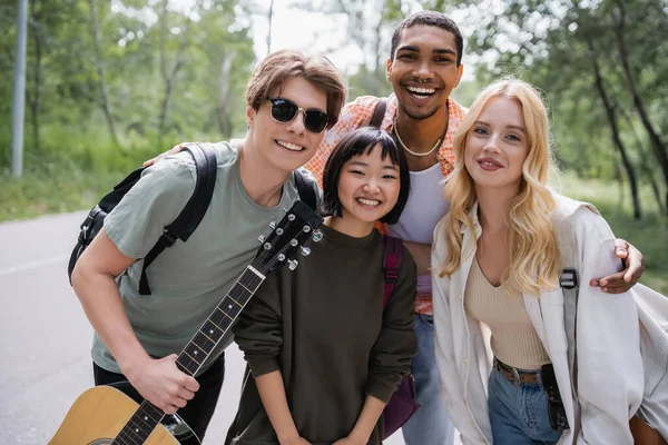 Man in sunglasses with guitar smiling at camera together with cheerful interracial friends - foto de stock