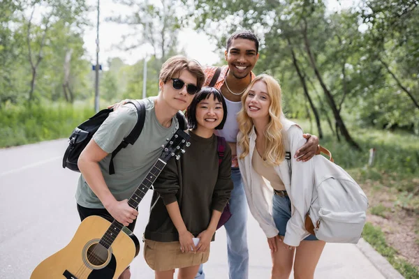 Joyful multicultural friends with backpacks and guitar smiling at camera on countryside road - foto de stock