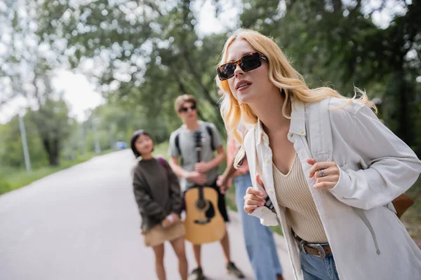 Blonde woman in sunglasses near multicultural friends on blurred background — Fotografia de Stock