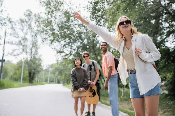 Woman in sunglasses stopping car near multiethnic friends on road in countryside — Foto stock