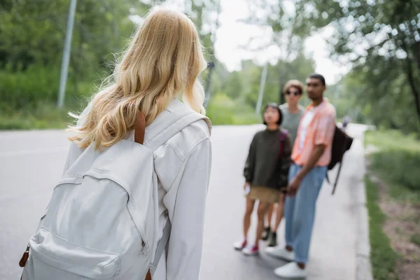 Blonde woman with backpack near blurred multicultural friends on countryside road - foto de stock