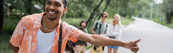 Smiling african american man stopping car with thumb up near hitchhikers on blurred background, banner — Stock Photo