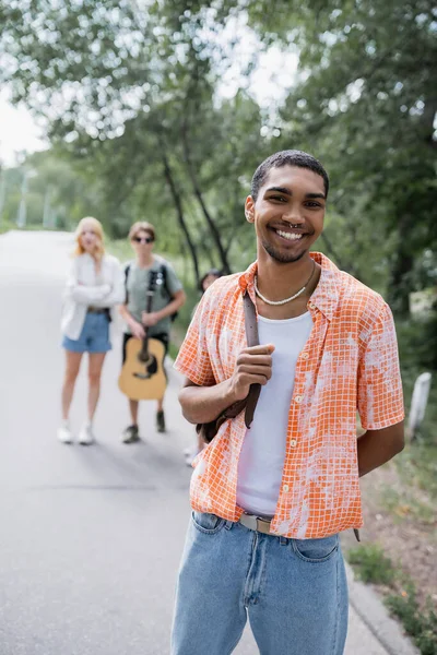 Happy african american traveler looking at camera near friends on blurred road — Stock Photo