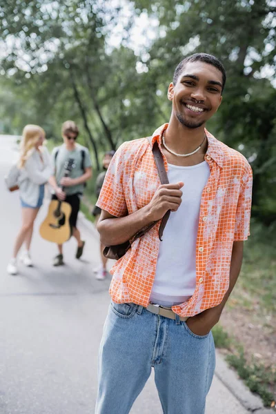 Happy african american man with hand in pocket looking at camera near friends on blurred road — Photo de stock