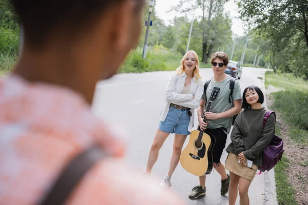 Happy interracial friends with guitar and backpacks looking at african american man on blurred foreground — Stock Photo