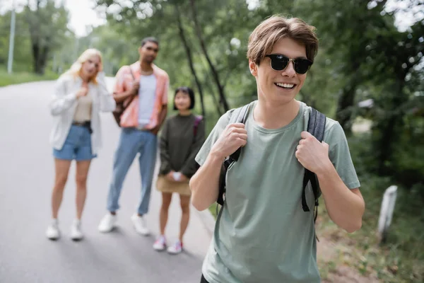 Молодий чоловік в сонцезахисних окулярах посміхається біля багатоетнічних друзів на розмитому фоні — стокове фото