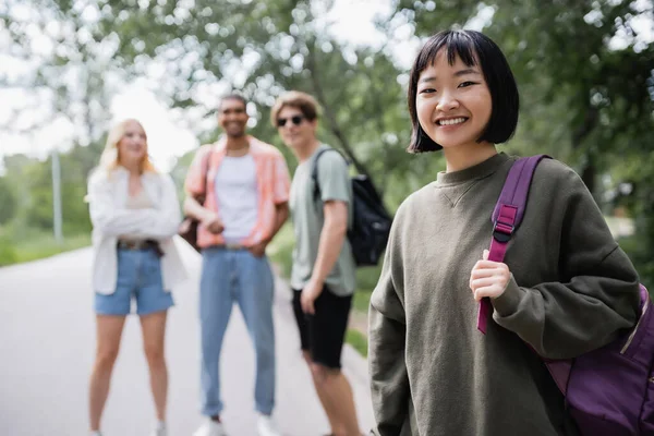 Brunette asian woman with backpack looking at camera near blurred multicultural friends — Photo de stock