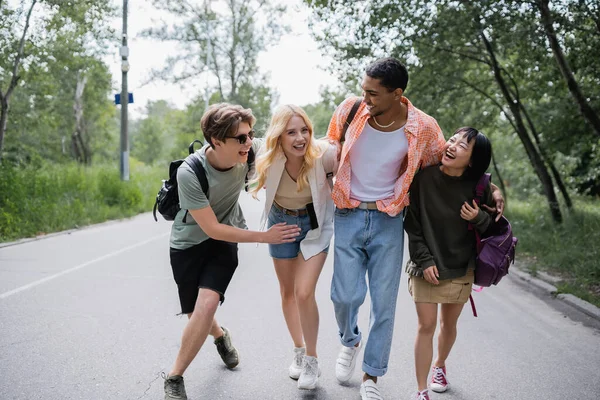 Full length of multiethnic tourists embracing and laughing during walk on countryside road - foto de stock