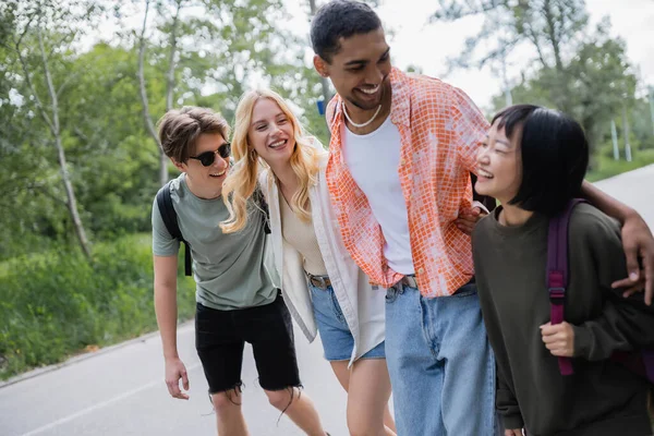Cheerful multiethnic travelers embracing and laughing during summer walk in countryside — Stock Photo