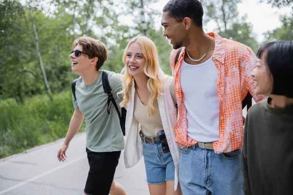 Cheerful multicultural travelers walking on road near forest - foto de stock