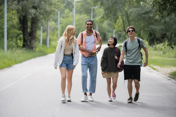 Full length of young interracial travelers with backpacks walking along road near forest — Foto stock