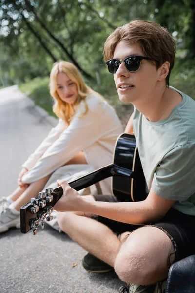 Young man in sunglasses playing acoustic guitar to blurred woman sitting on road — Stock Photo