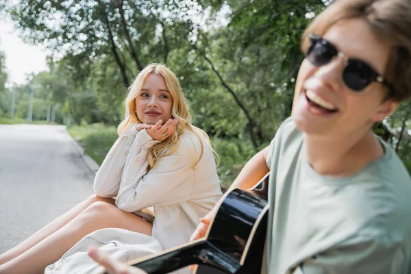 Blonde woman looking at happy man playing guitar on blurred foreground — Stockfoto