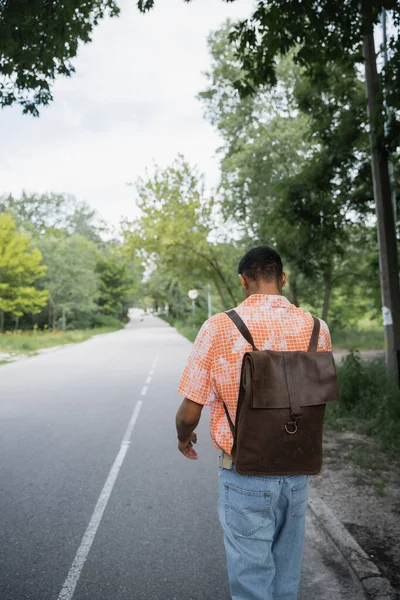 Назад вид на Африканский американский путешественник с кожаным рюкзаком ходьба по дороге — стоковое фото