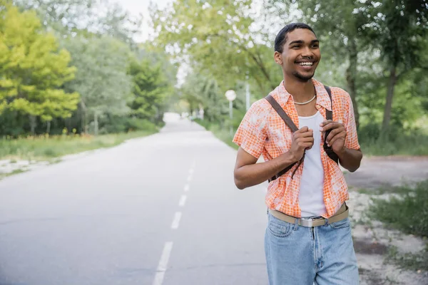 Happy african american traveler with backpack walking on road near forest — Photo de stock