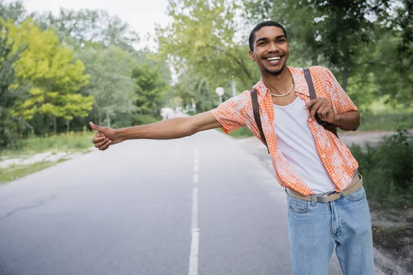 Joyful african american hitchhiker with thumb up stopping car on road in countryside - foto de stock