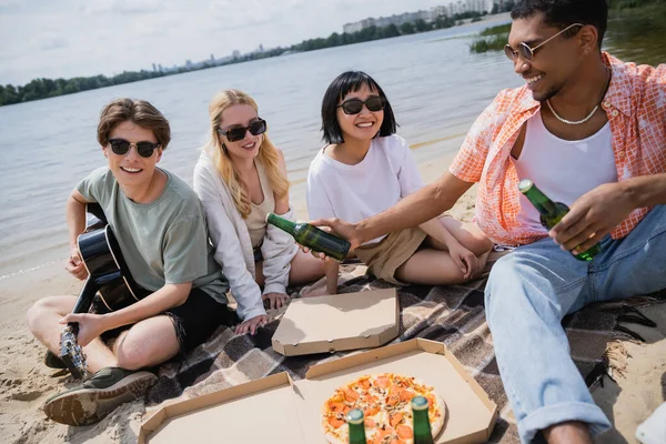 Smiling man in sunglasses playing guitar during beach party with multicultural friends — Foto stock