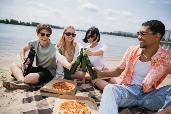 Happy interracial friends in sunglasses clinking beer bottles during beach party — Stock Photo