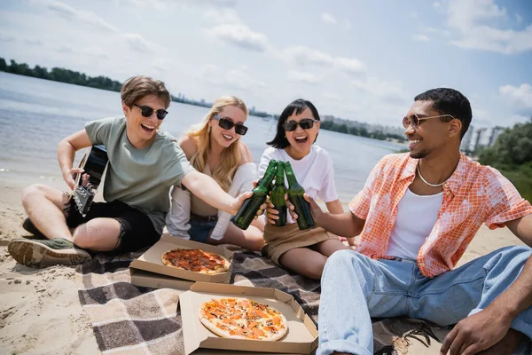 Happy multiethnic friends in sunglasses toasting with beer near pizza on blanket — Fotografia de Stock